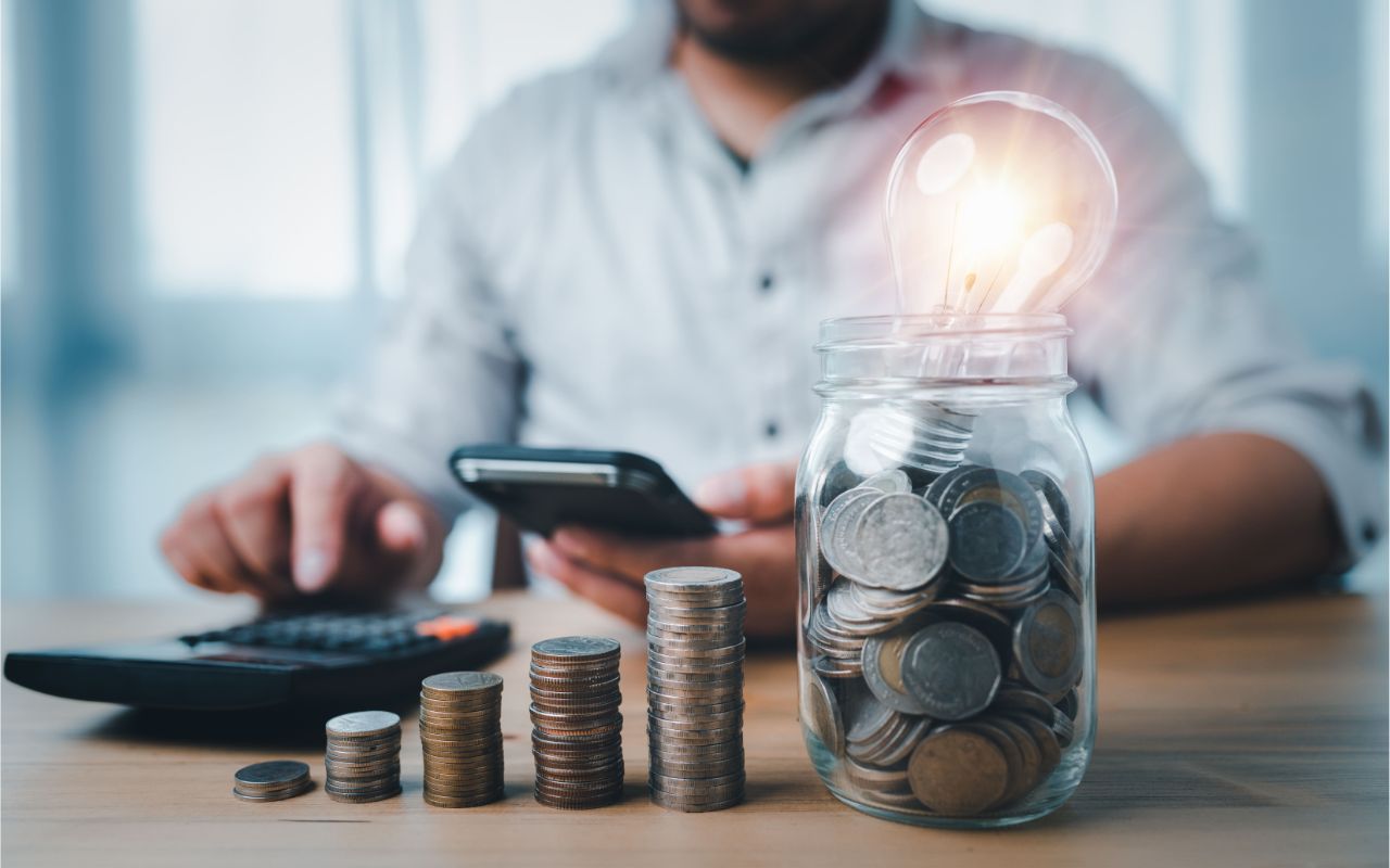 coins stacking on desk and lightbulb in jar with businessman use smartphone and calculator saving energy and money concept idea for save or investment